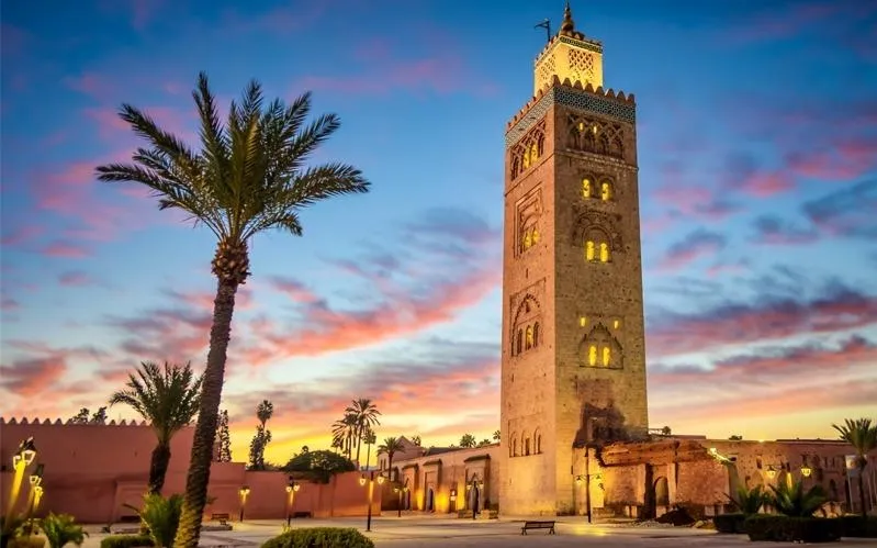Tour de l'horloge de la mosquée de Marrakech au coucher du soleil, illuminée par des teintes dorées et orangées.