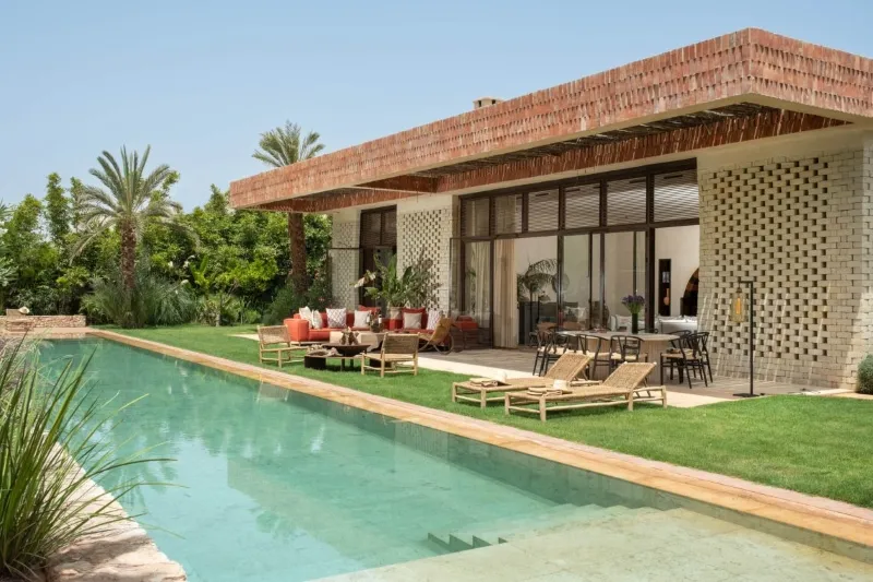 Vue de la piscine et de la terrasse d'une villa de luxe à Maroc Séjour, entourée de palmiers et de chaises longues.