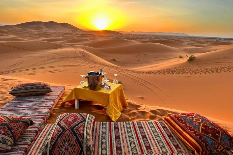 Un coucher de soleil sur le désert marocain, avec une table et des coussins disposés pour se détendre.