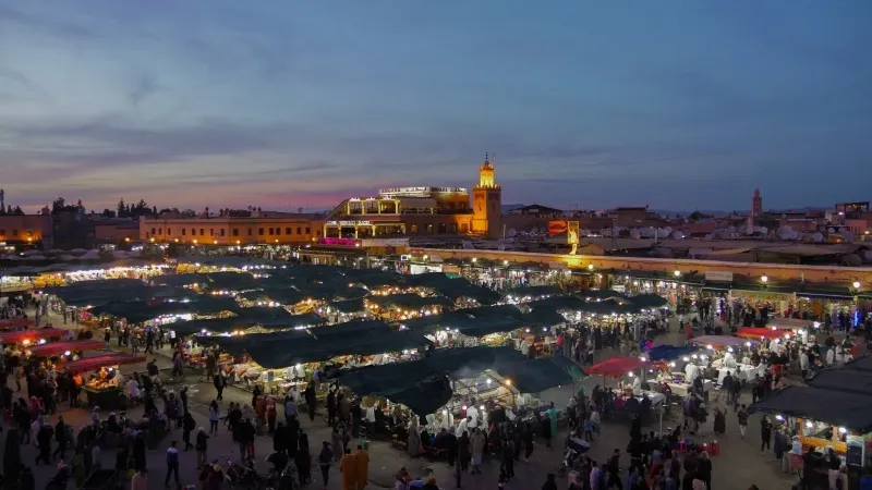 Marché de Marrakech au crépuscule, illuminé par des lanternes, avec des silhouettes de parapentes dans le ciel.