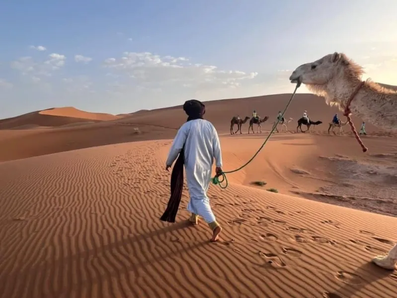 Un homme marche avec un chameau dans le désert marocain, entouré de dunes de sable sous un ciel ensoleillé.