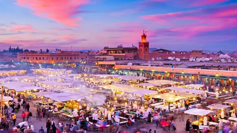 Marrakech au crépuscule, illuminée par des lumières chaudes, avec des silhouettes de bâtiments emblématiques en arrière-plan.